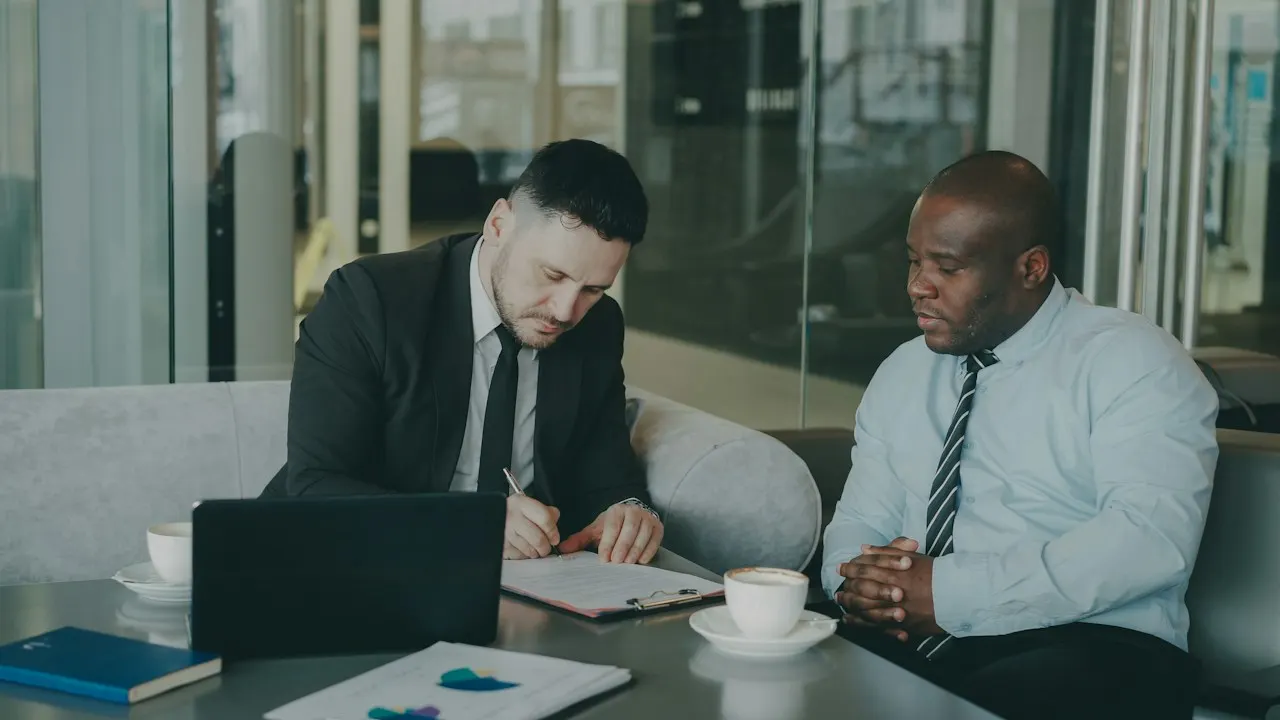 Two businessmen at a table, one signing a document such as a management agreement