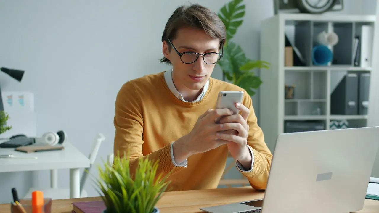 A man at a desk checking a payment on his phone next to a laptop